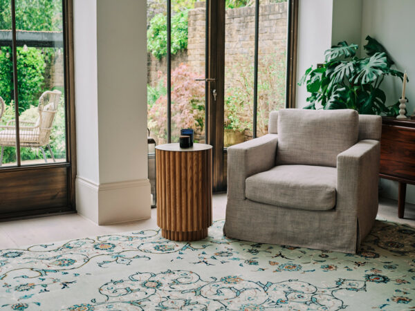 A living room with a London Persian Rug Company carpet, grey armchair, side table, and garden views through large windows.